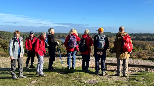 Eight people in walking attire standing at the top of a hill admiring a view. Five people have their backs to the camera and three people are looking at the camera.
